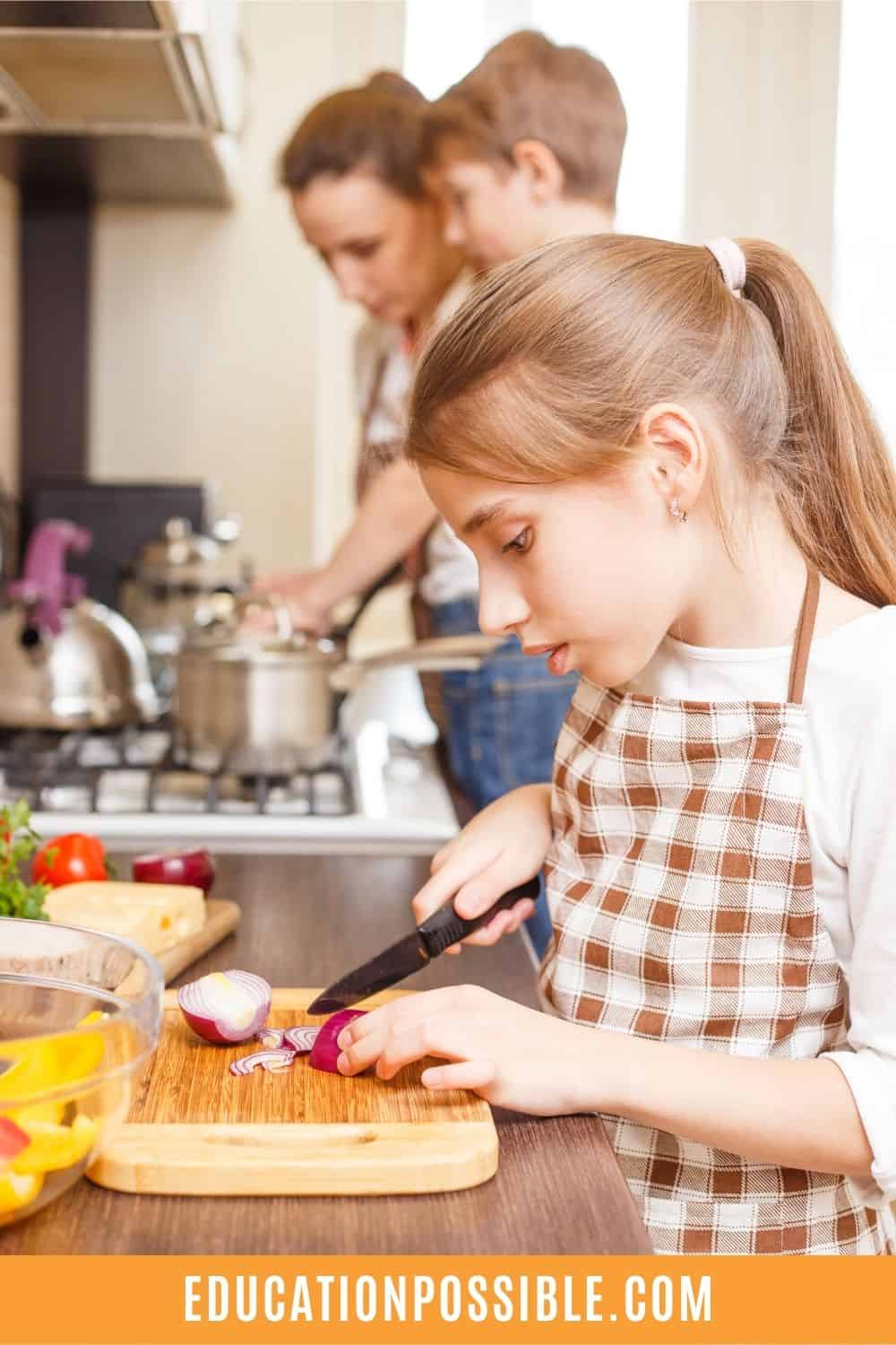 Tween girl cutting an onion on a cutting board while her mom and brother make something on the stove.