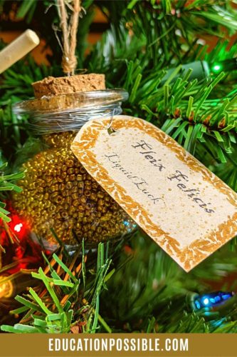 Small glass jar ornament filled with small gold beads, with a beige tag, hanging on a Christmas tree.