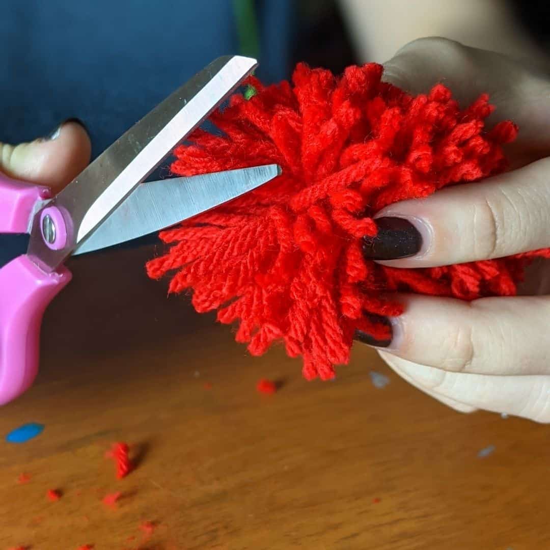 Girl using scissors to trim the ends off a red yarn pom-pom