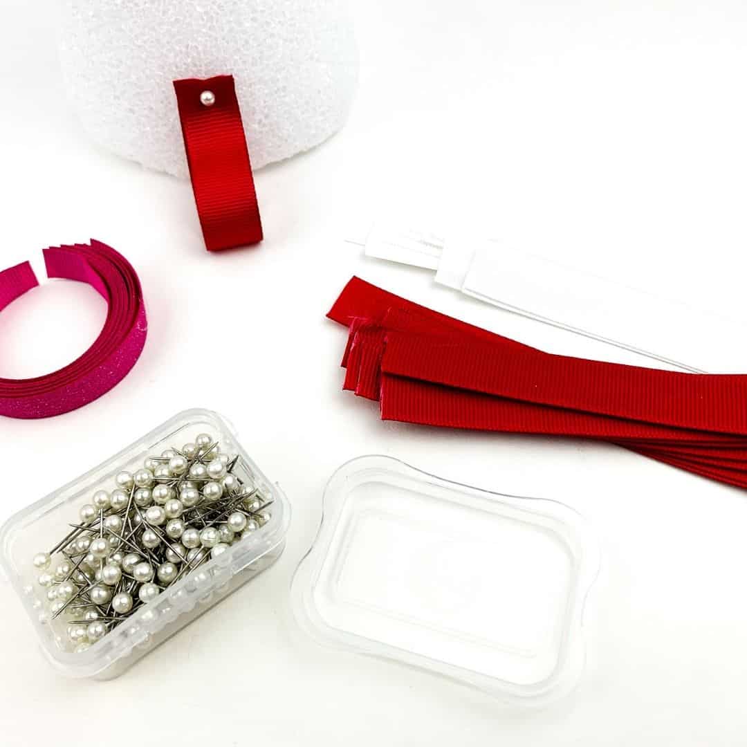 Strips of red, white, and pink ribbon and pearl pins. A red ribbon loop is pinned onto the bottom of a Styrofoam cone, for a ribbon tree craft.