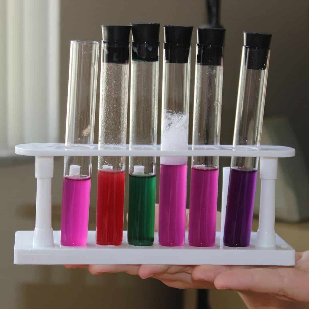 6 test tubes in a white holder with various colored liquids inside. Girl holding the tray in her hands.