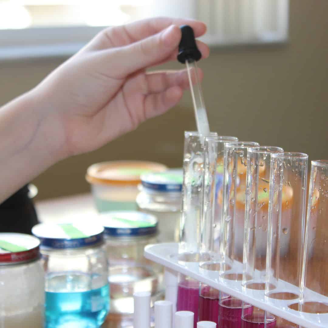 Tween girl holding an eye dropper with cloudy liquid in it and adding it to a row of test tubes with purple liquid inside.