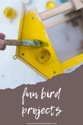 Hand holding a paintbrush, painting a wooden birdhouse yellow.