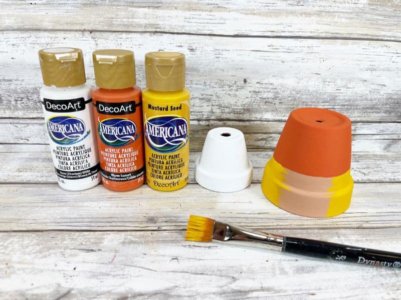 Bottles of paint, white, yellow, and orange being used to paint a clay pot like a candy corn