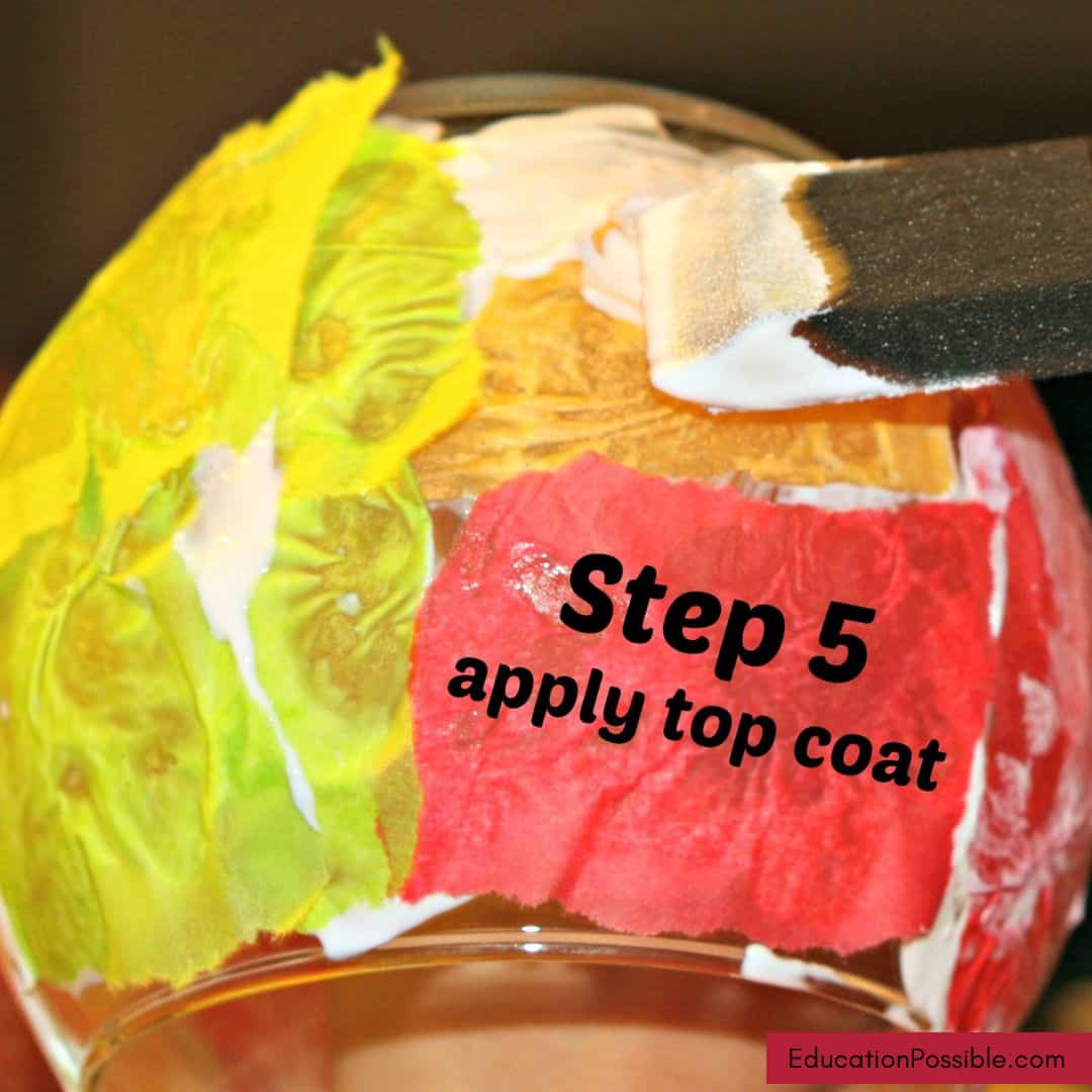 Using a foam brush to add a top coat of Mod Podge over tissue paper on a glass candle holder.