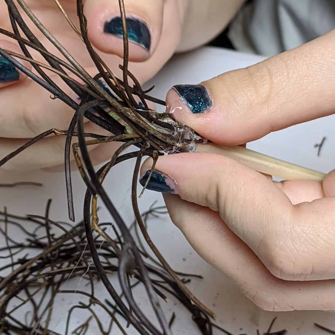 Girl gluing thin twigs to the end of a chopstick to make a mini broom