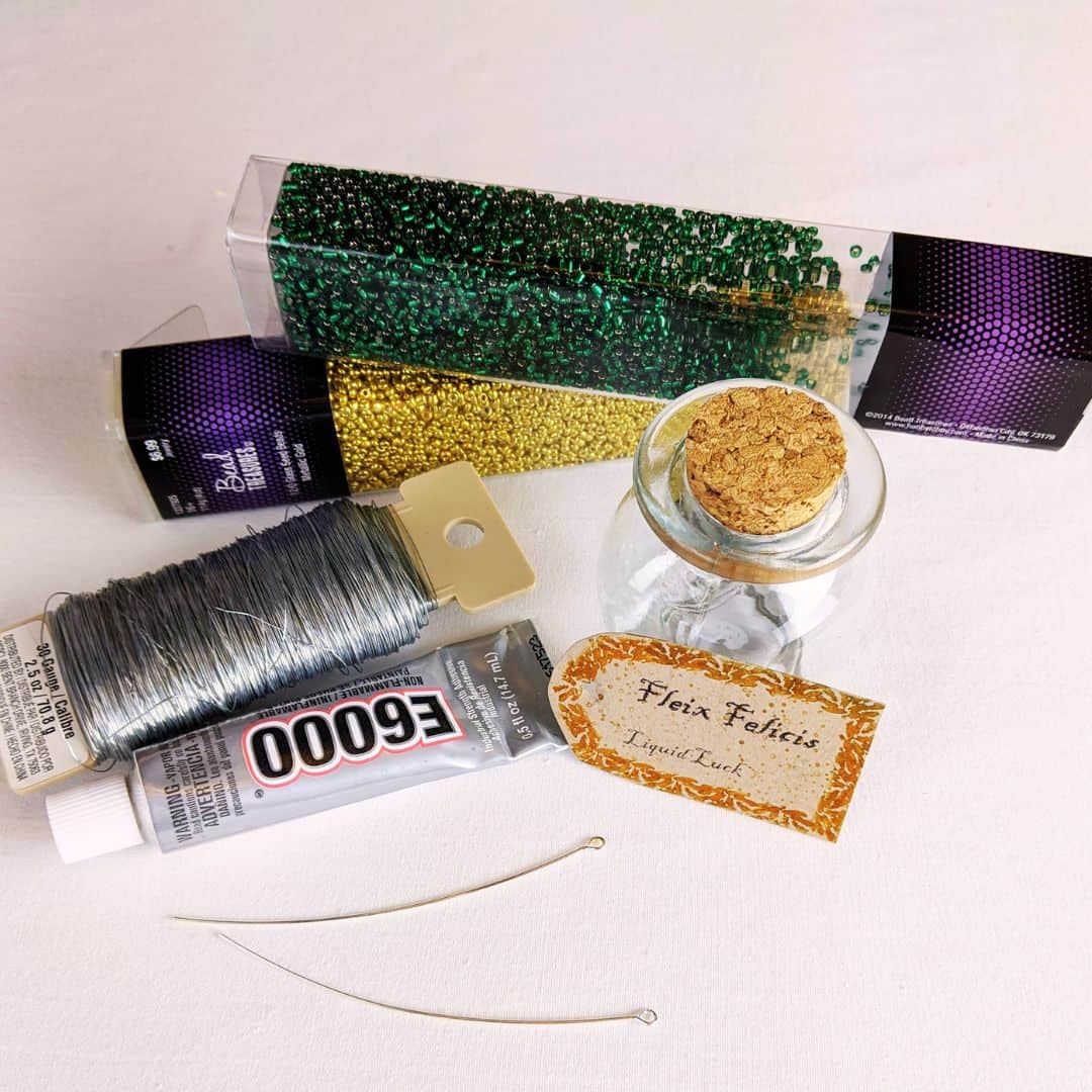Materials needed to make a Harry Potter potion ornament. Green and gold glass beads, jewelry wire, glue, eye pins, a tag, and a glass jar.