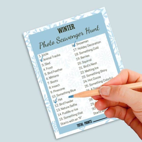 Printable winter-themed scavenger hunt being checked off by a mock-up hand holding a blue lead pencil.