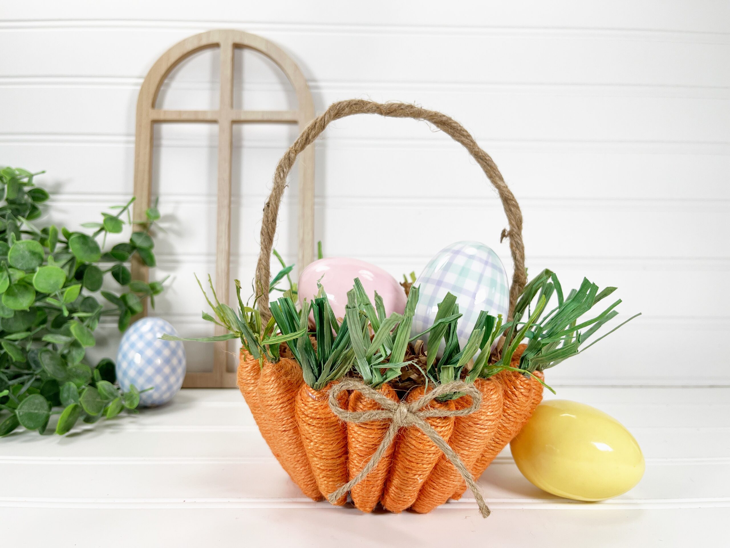 Twine wrapped carrots wrapped around a cup with a jute handle - a DIY Easter basket. It sits on a white table with colorful and plaid plastic Easter eggs inside and next to it.