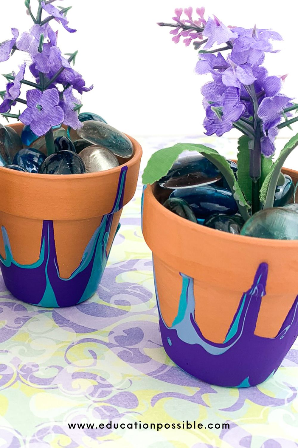 Two DIY pour paint flower pots on a purple/green patterned fabric. Pots filled with light purple silk flowers and glass beads.