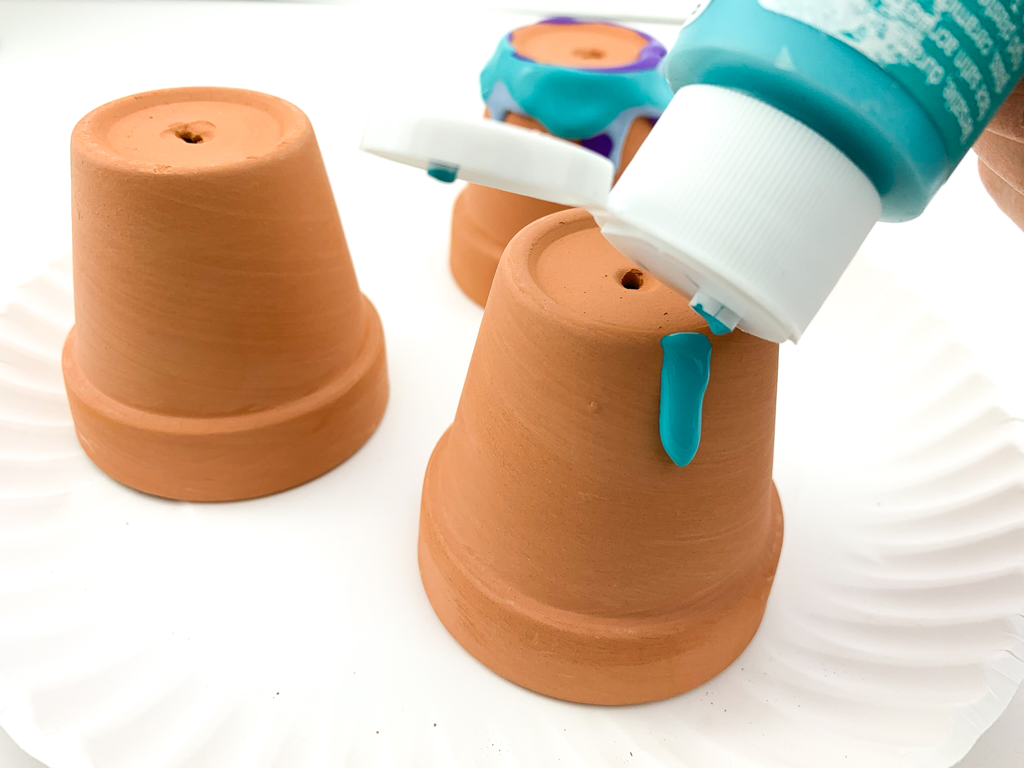 3 mini terracotta pots upside down on a paper plate. One is plain and one has 2 colors of blue and purple paint running down from the bottom. Someone is pouring blue paint from a bottle onto the third pot's bottom.